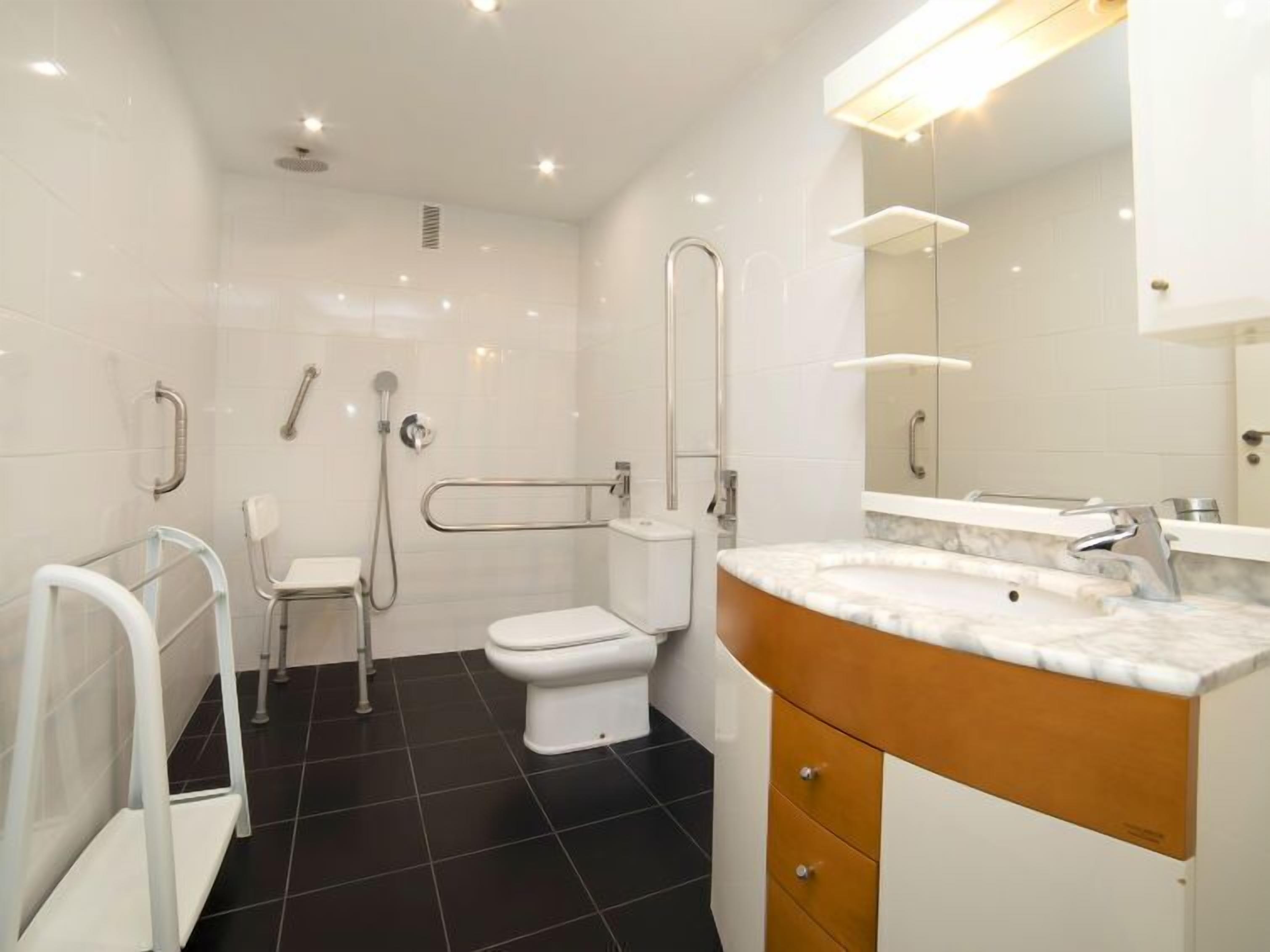 A modern bathroom with a white toilet, a shower with grab bars, a vanity with a marble countertop, and a large mirror.