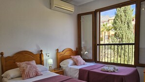Habitación de hotel con dos camas, cada una con cabecero de madera, una ventana con vista a edificios y árboles, y un equipo de aire acondicionado en la pared.
