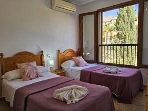 Hotelkamer met twee bedden, elk met een houten hoofdeinde, een raam met uitzicht op gebouwen en bomen, en een airconditioningsunit aan de muur.