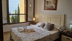 Habitación de hotel con cama, mesita de noche, lámpara, un cuadro y ventana con vistas a árboles y edificios.