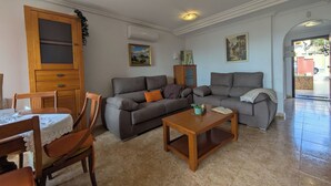 Una sala de estar con un sofá gris, sillas de madera, una mesa de centro de madera y una mesa de comedor con sillas. Hay un aparador de madera y un cuadro en la pared.