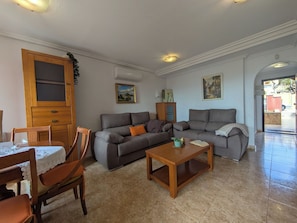 A living room with a grey sofa, wooden chairs, a wooden coffee table, and a dining table with chairs. There is a wooden cabinet and a painting on the wall.