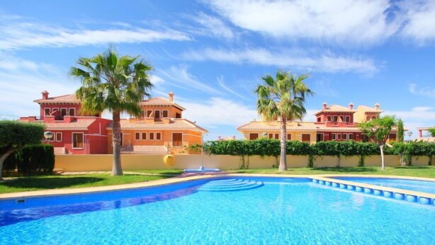 Una piscina con superficie de agua azul cristalina, rodeada por dos casas grandes con fachadas rojas y amarillas, y palmeras.
