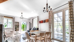 A spacious living area with a dining table, chairs, and a chandelier. There is a sofa and a TV in the background.