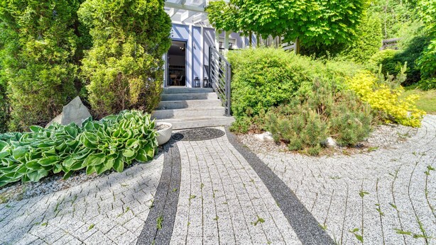 A house with a paved driveway leading to a front door, surrounded by lush greenery and a well-maintained garden.