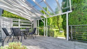 A covered outdoor patio with a table and chairs, surrounded by a white fence and lush greenery.