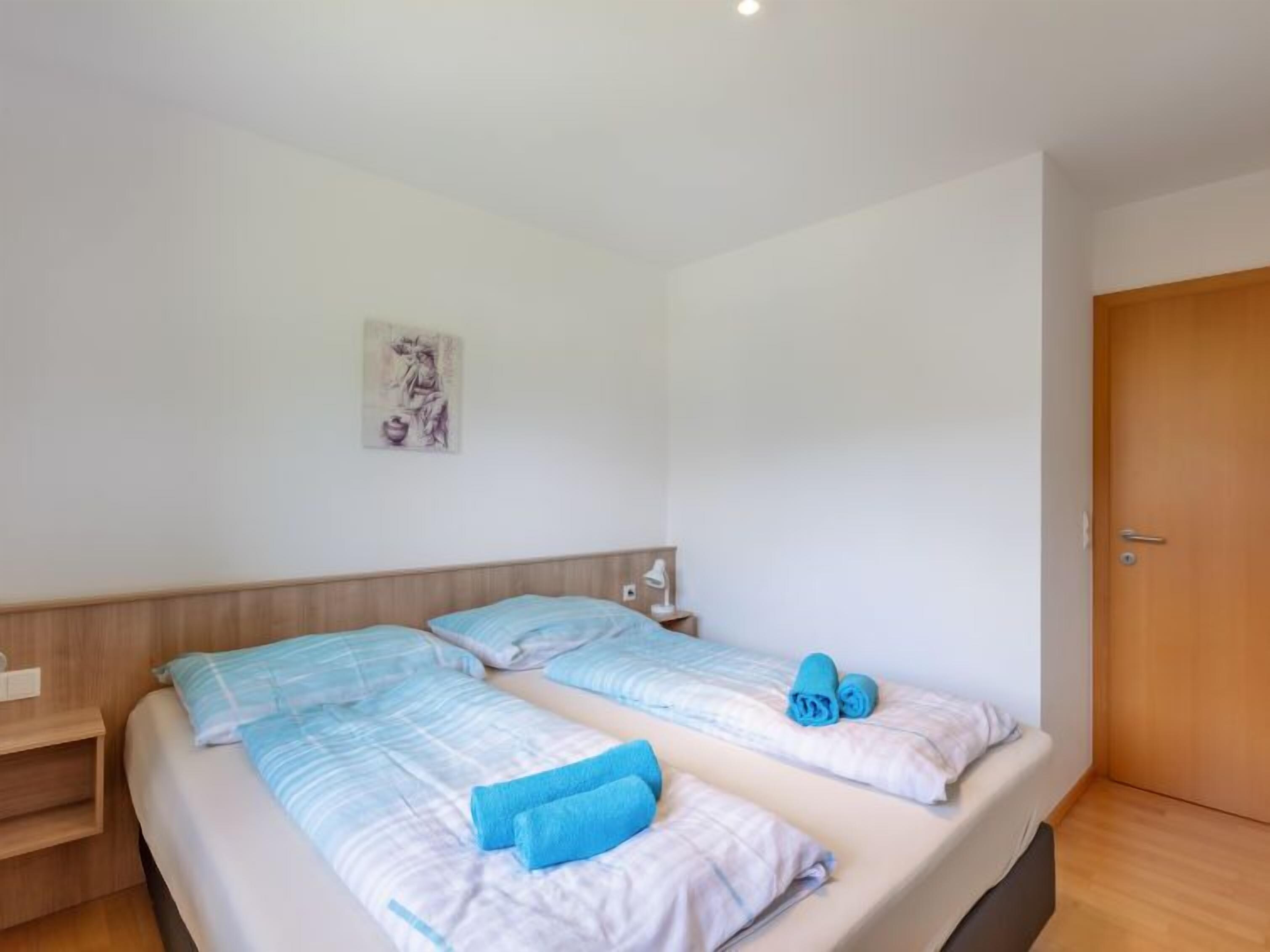 Una habitación con dos camas, cada una con ropa de cama y almohadas azules, una puerta de madera y una mesita con una lámpara.