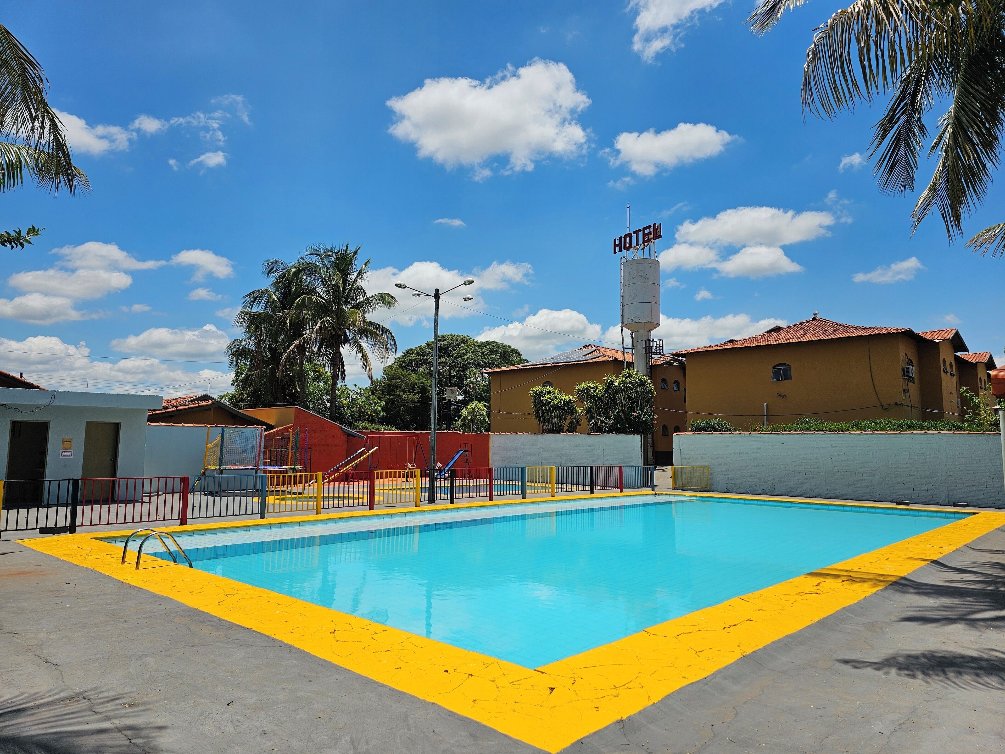 A swimming pool with a yellow safety border, a sign indicating a hotel, and a red-roofed building in the background.