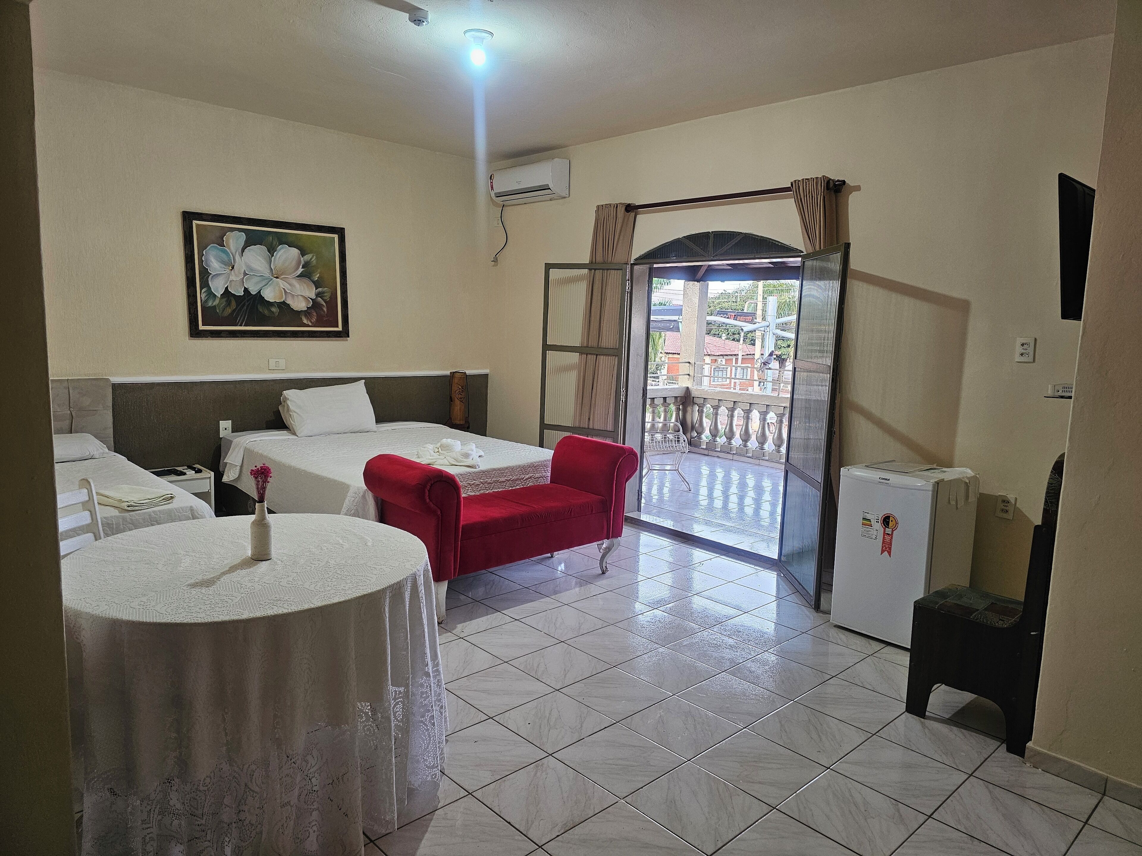 Habitación de hotel con dos camas, una silla roja, una mesita con un florero y un balcón con vistas.