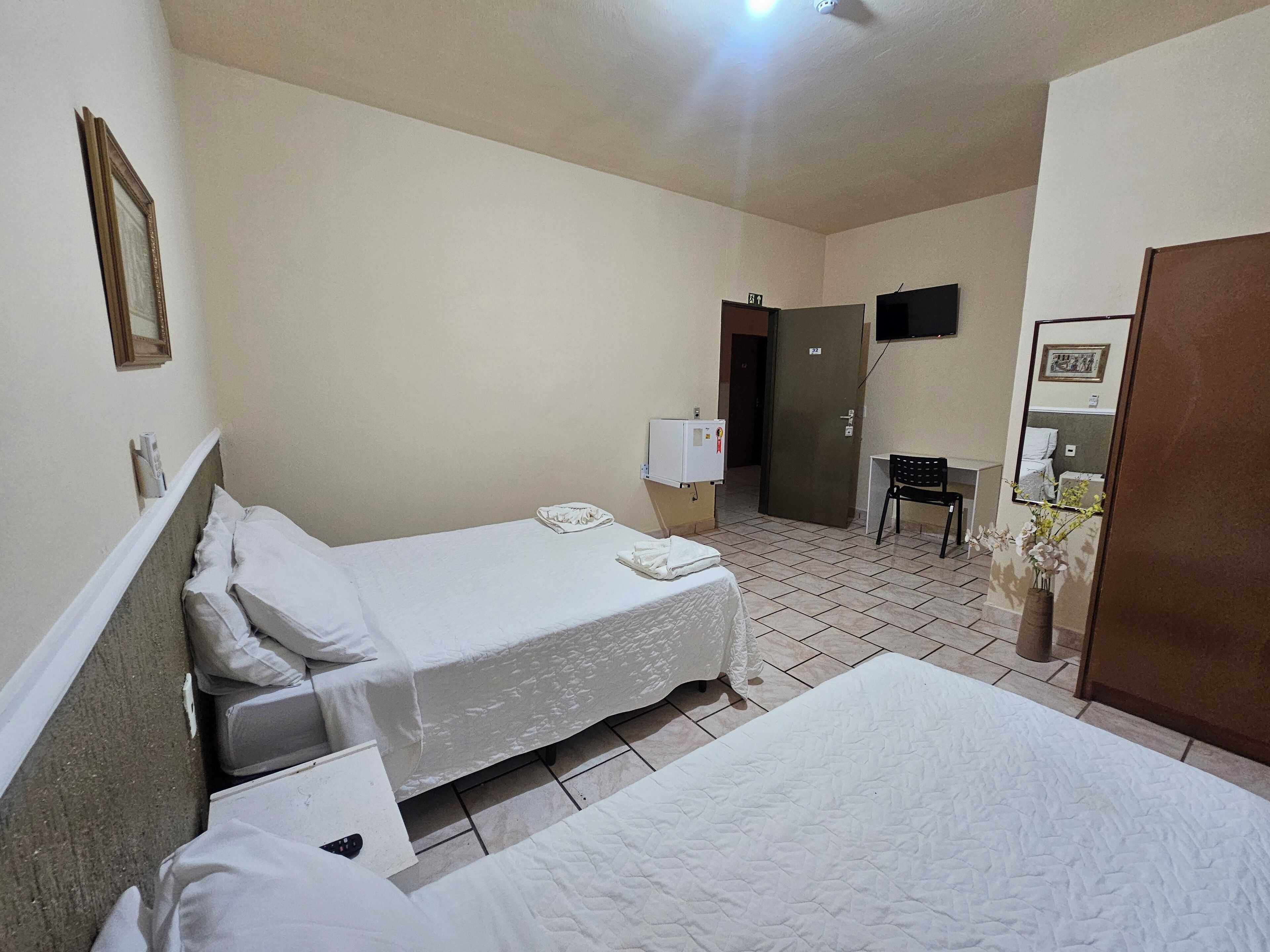 Une chambre d’hôtel avec deux lits simples, un bureau avec une chaise, une petite armoire, un téléviseur fixé au mur et une porte donnant sur une autre pièce.