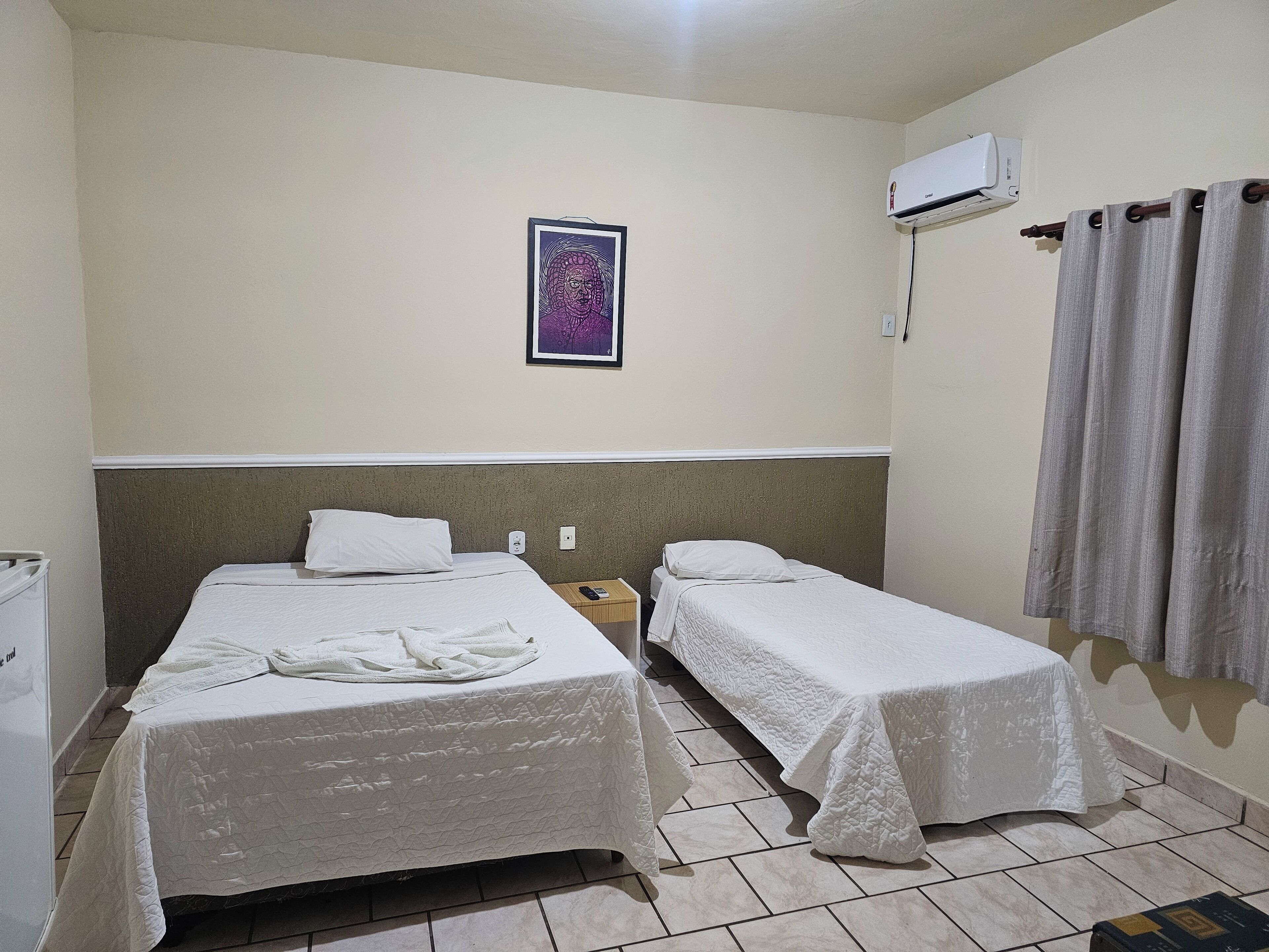 A hotel room with two single beds, a small bedside table with a phone, an air conditioning unit, and a framed artwork on the wall.