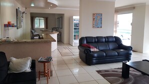A living room with a black leather sofa, a wooden coffee table, and a small wooden stool. There is a kitchen area in the background with a refrigerator and a dining table. - Esplanada Hotel (Paraguacu Paulista)