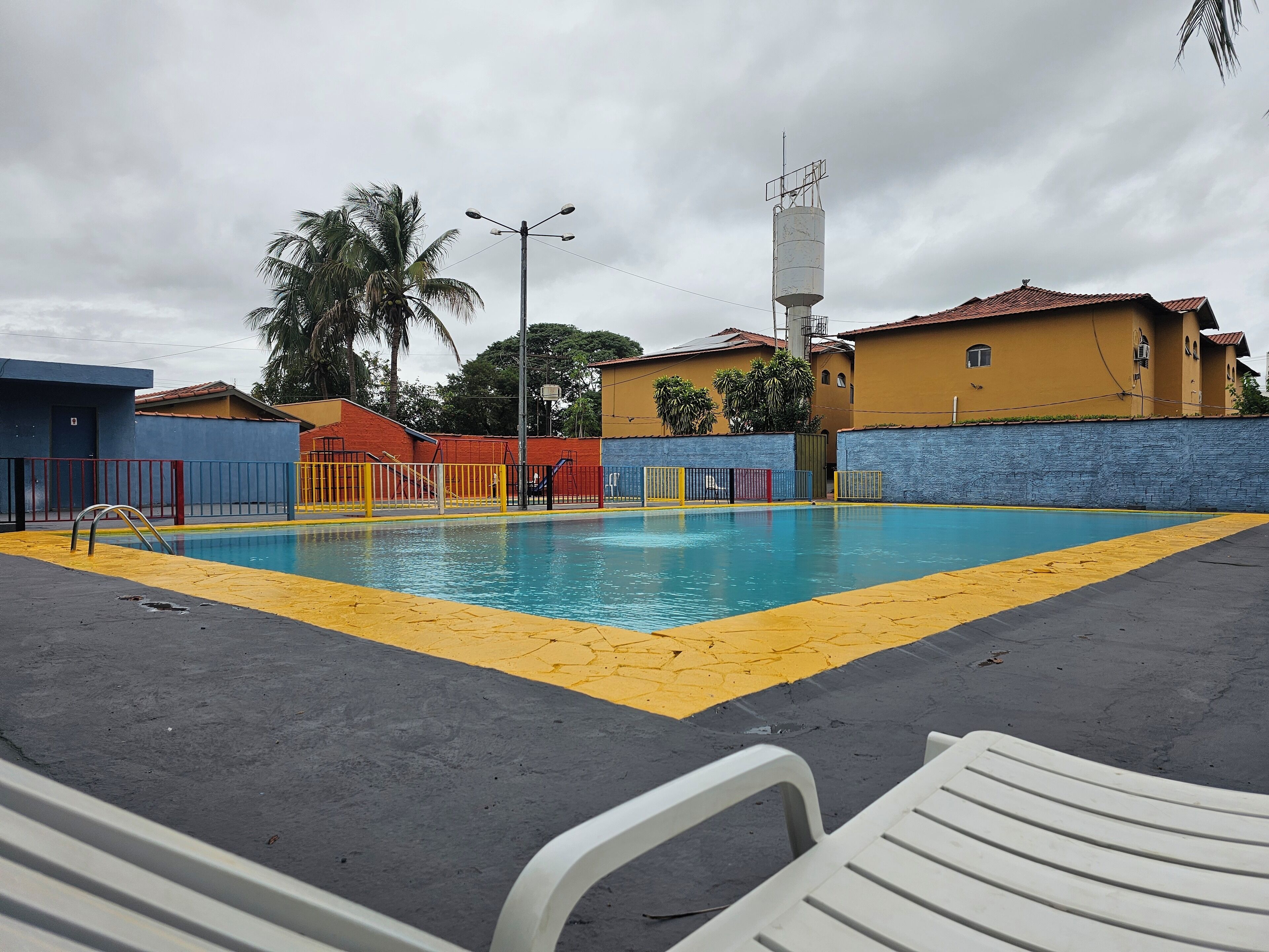 Ein Schwimmbecken mit gelber Sicherheitslinie, umgeben von einem niedrigen Zaun. Im Hintergrund sind Gebäude mit roten und gelben Wänden zu sehen.