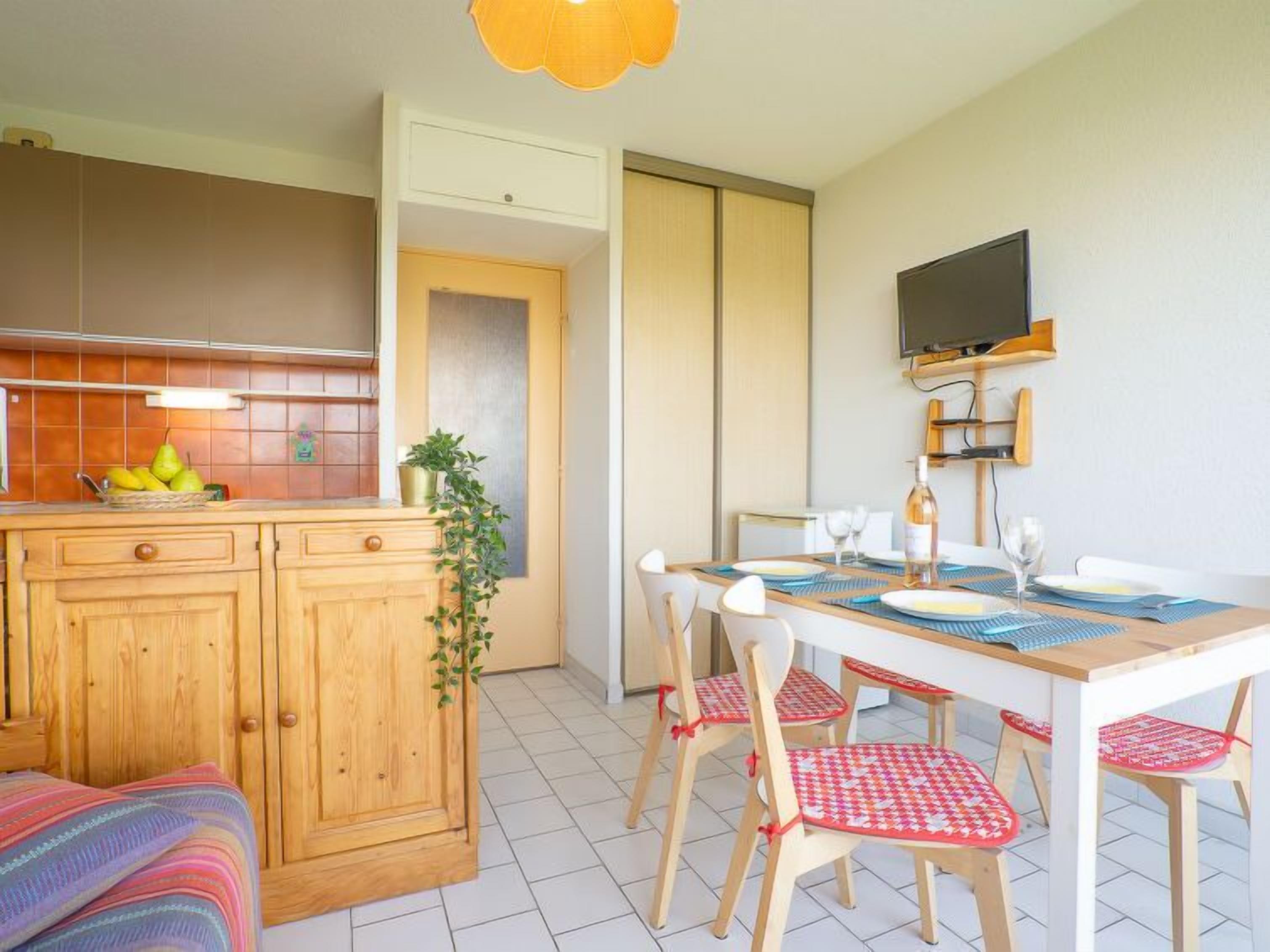 Una cocina compacta con armarios de madera, una mesa de comedor con sillas y un televisor montado en la pared.