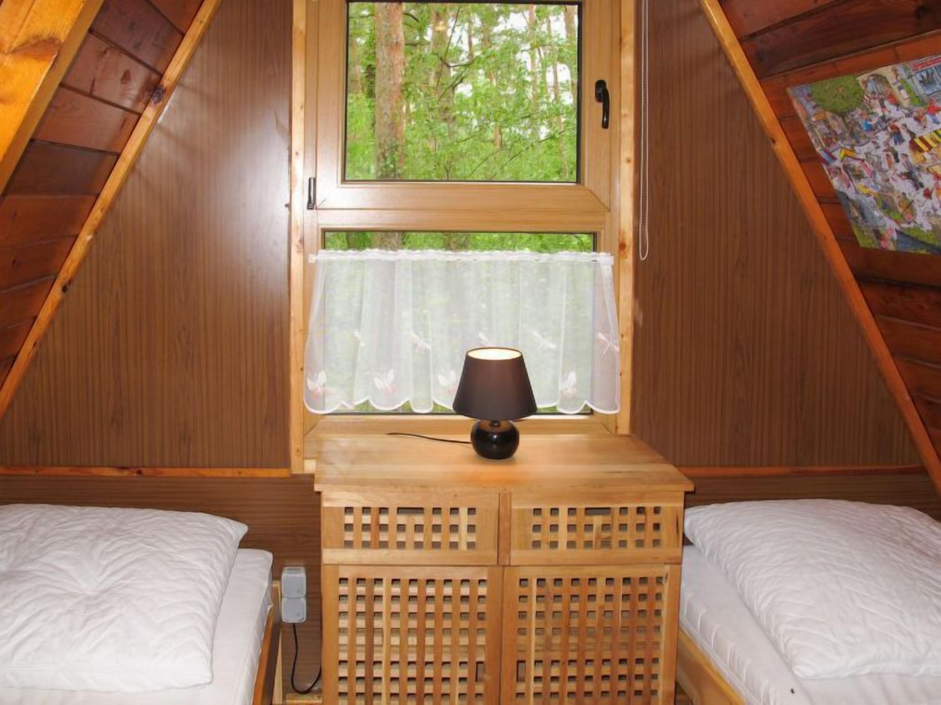 Un dormitorio acogedor en el ático, con techo inclinado de madera, una ventana con cortina translúcida, una lámpara de noche y dos camas de madera.