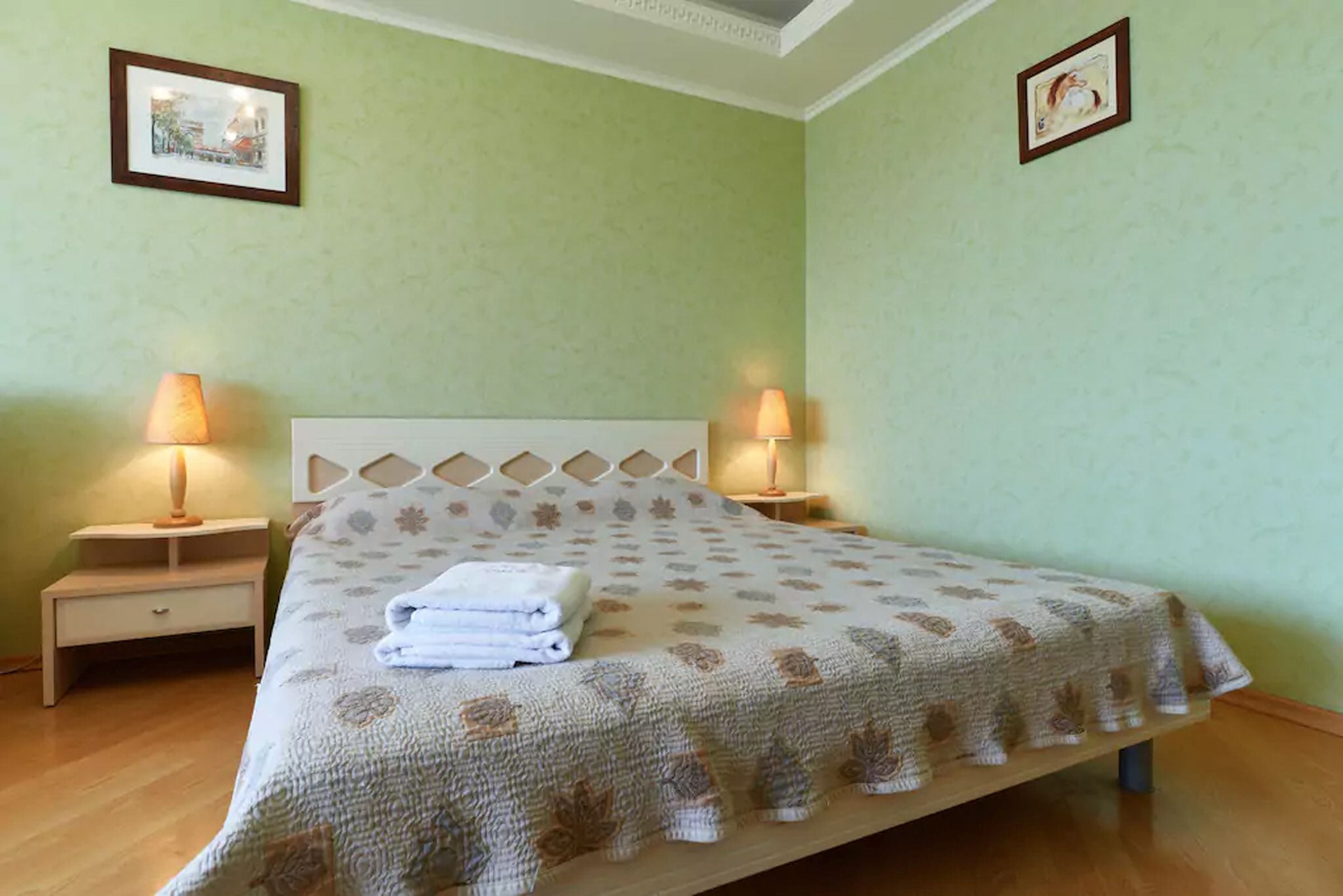 Una cama bien hecha con una colcha estampada, dos lámparas de noche y una mesita de noche con cajón.
