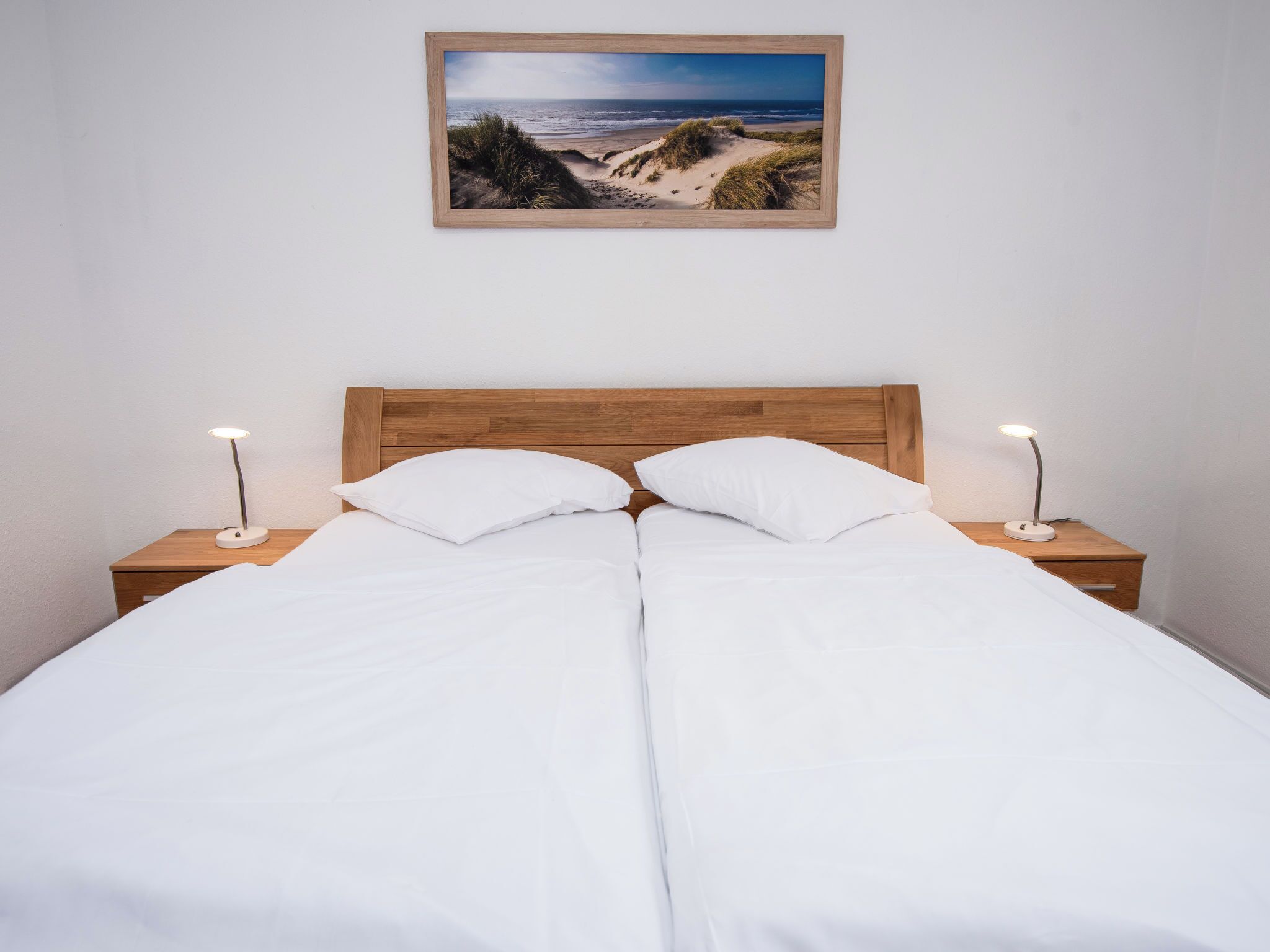 A double bed with white linens, two bedside tables with lamps, and a framed beach landscape picture on the wall.