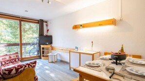 A cozy, well-lit room with a dining table set for two, a television, and a bicycle outside. - Lunik Orion by Interhome (Le Corbier)