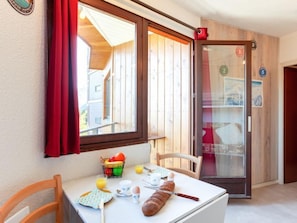Un pequeño comedor con una mesa lista para el desayuno, una ventana con cortina roja y una puerta de madera con indicaciones numéricas.