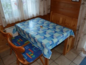 Ein Essbereich mit Tisch, der mit einer blau-weißen Tischdecke bedeckt ist, Holzstühle mit bunten Kissen und eine Holzvitrine an der Wand.