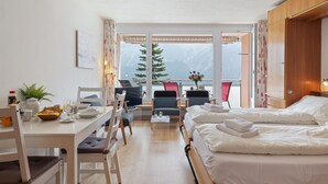 Una camera d'albergo con un letto, un tavolo da pranzo con sedie, una TV e vista sulle montagne attraverso ampie finestre.