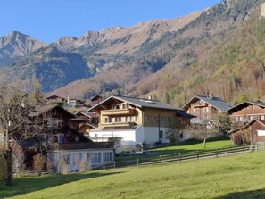 Un pueblo de montaña con casas de madera tradicionales, un primer plano con césped y montañas escarpadas al fondo.