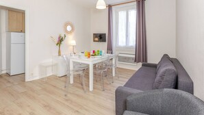 A modern living room with a grey sofa, a dining table with clear chairs, a white refrigerator, and a window with curtains.
