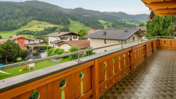 A balcony with a wooden railing overlooking a residential area with houses and green hills in the background.