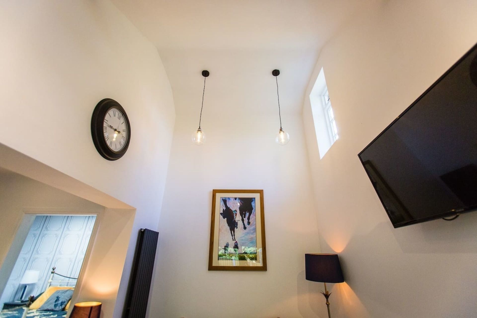 A modern hotel room with a large bed, a mounted TV, a framed artwork, and a ceiling light fixture.