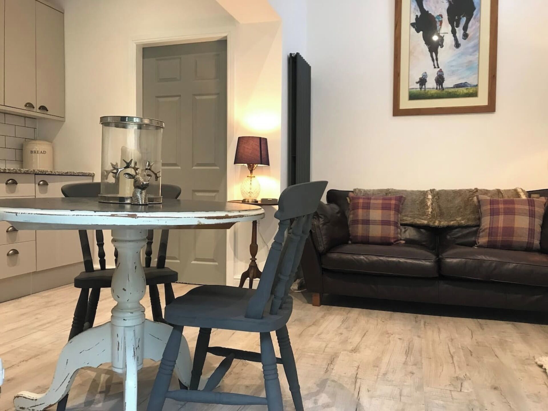 A dining area with a round table and chairs, a kitchen in the background, a sofa with plaid cushions, and a framed picture on the wall.