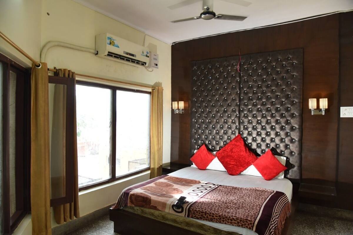 A hotel room with a bed, a window with curtains, an air conditioning unit, a ceiling fan, and a wall-mounted light fixture.