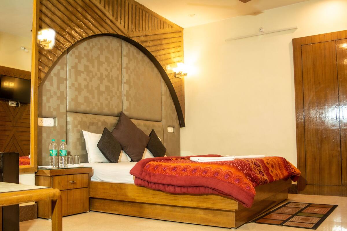 A hotel room with a wooden headboard, a bed with a red blanket, a wooden nightstand, a chair, and a television on the wall.