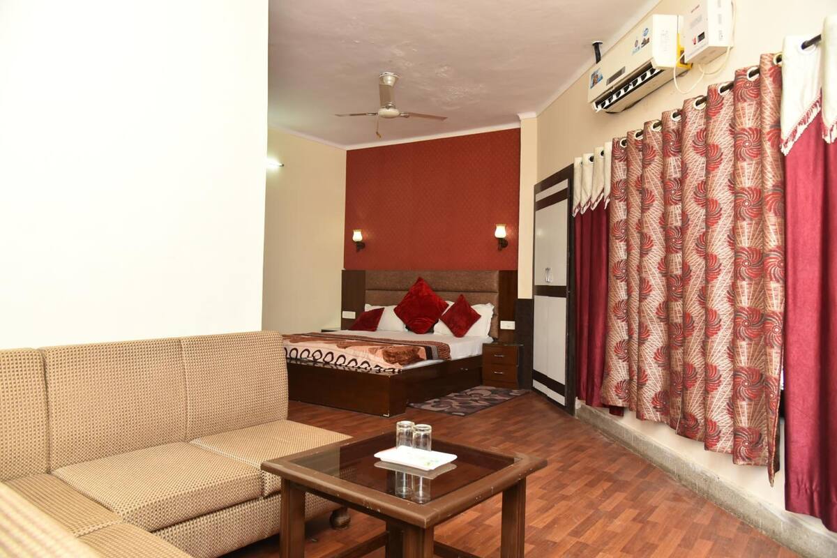 A hotel room with a sofa, a bed with red pillows, a ceiling fan, and patterned curtains.