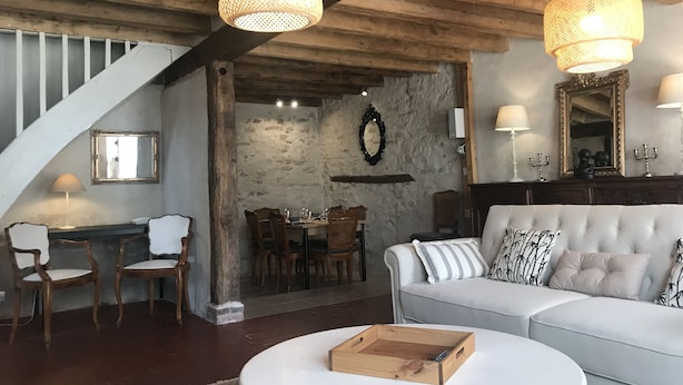 Una sala de estar acogedora con una escalera, vigas de techo de madera, un comedor y un sofá blanco con cojines estampados.
