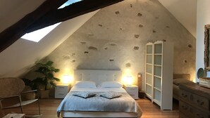 Un dormitorio con techo inclinado, una cama con colcha estampada, lámparas de noche, una mesita de noche, un aparador de madera y una planta en maceta.