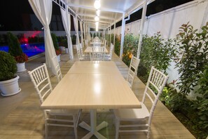 An outdoor covered patio with a long table and chairs, surrounded by potted plants and a pool. - Gardenia Hotel & Spa (Veles)