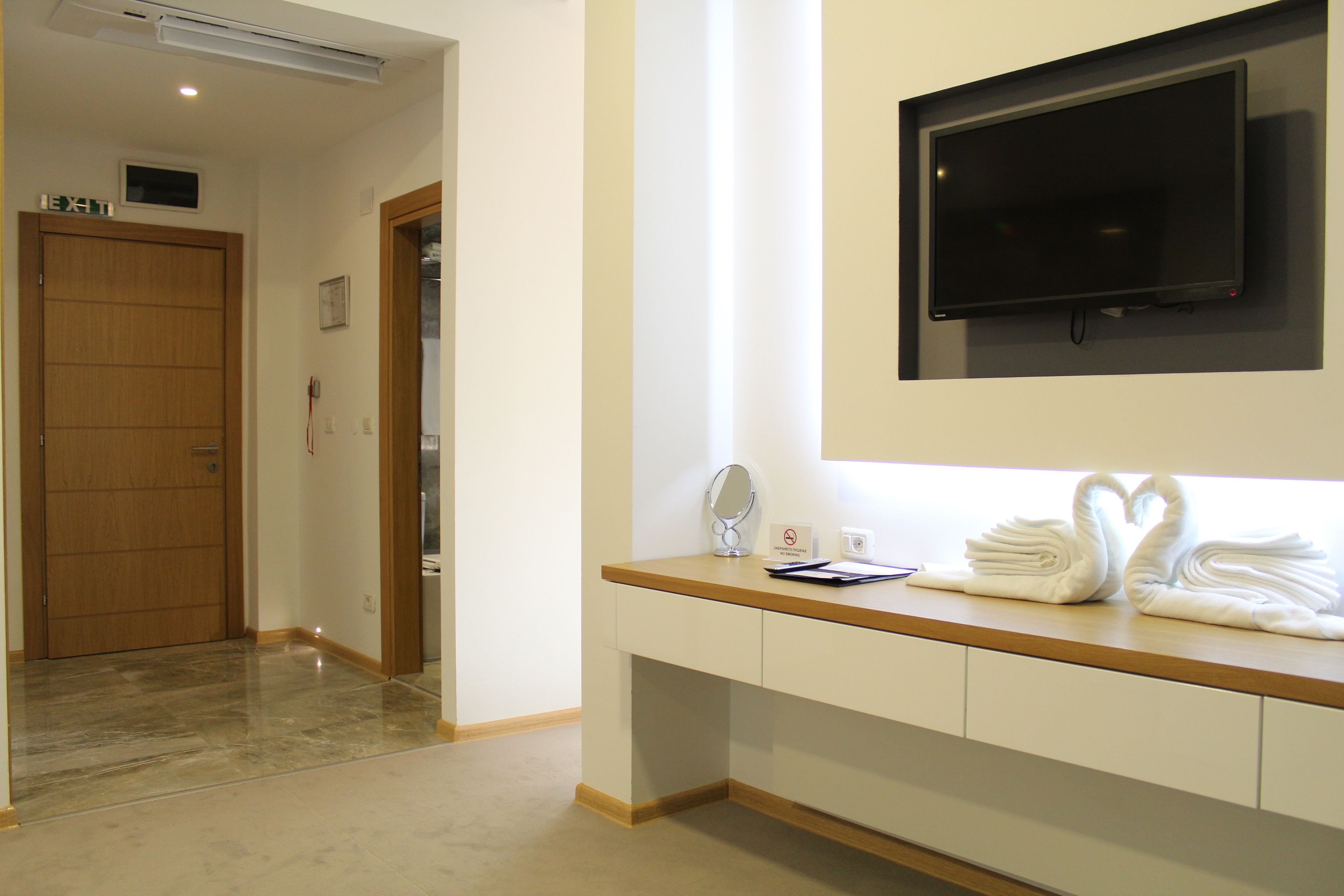 Habitación de hotel con puerta de madera, televisor de pantalla plana montado en la pared, tocador con espejo y toallero con toallas dobladas.