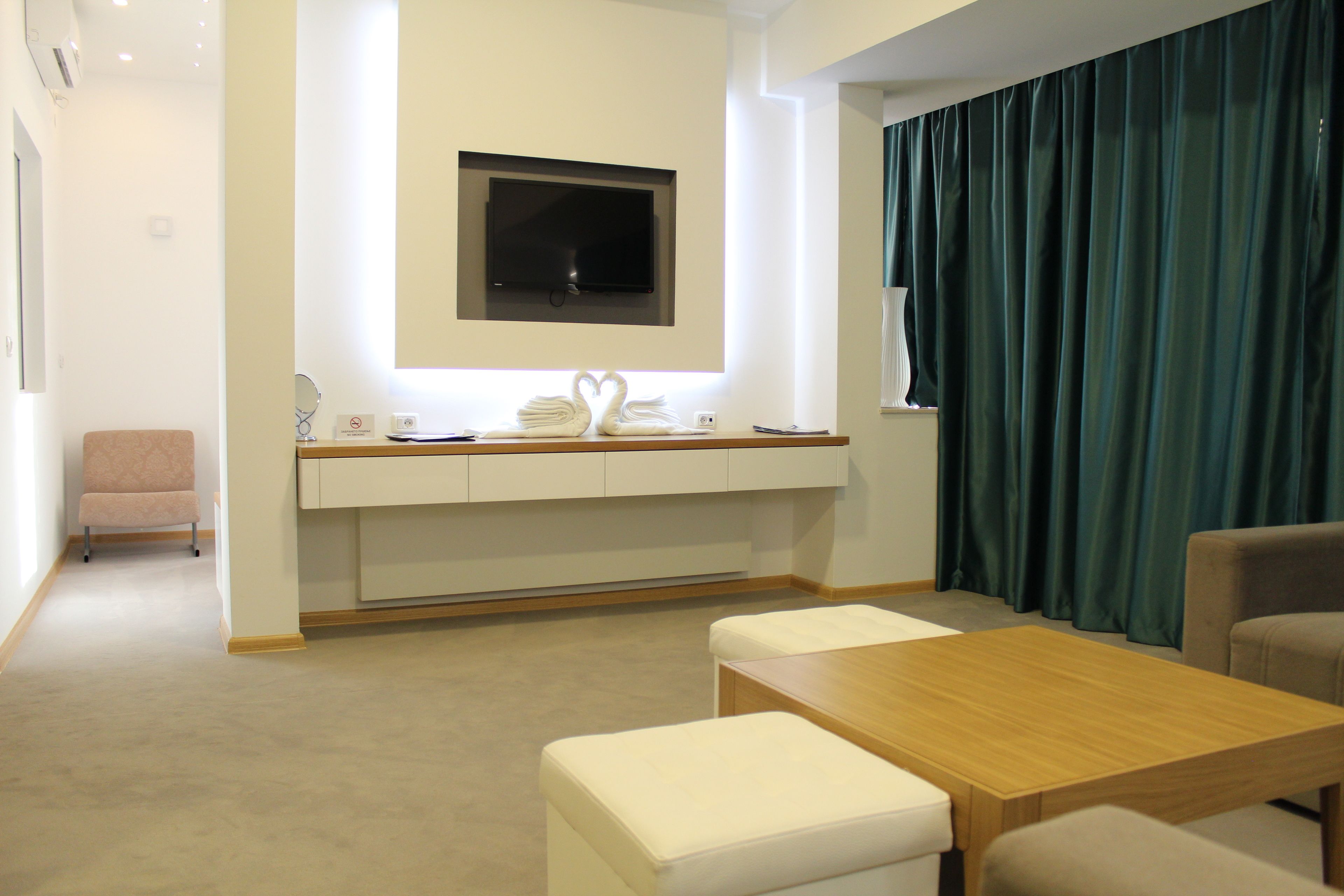 Ein modernes Hotelzimmer mit einem Holztisch, weiß gepolsterten Sitzgelegenheiten, einem flachbildfernseher an der Wand und einem dunkelgrünen Vorhang.