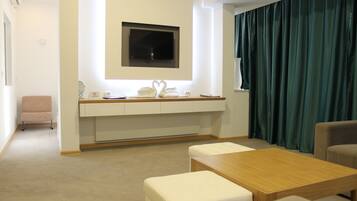 Ein modernes Hotelzimmer mit einem Holztisch, weiß gepolsterten Sitzgelegenheiten, einem flachbildfernseher an der Wand und einem dunkelgrünen Vorhang.