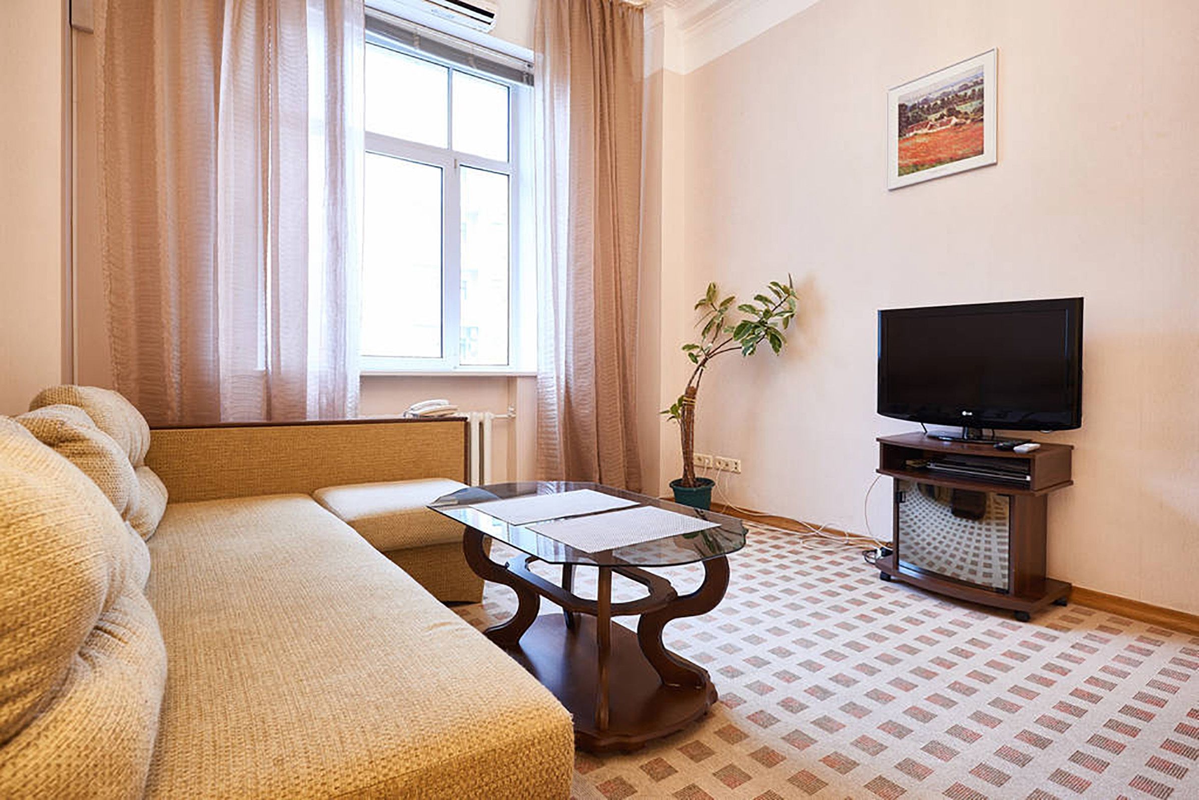 A living room with a sofa, a glass-top coffee table, a television on a stand, a plant, and a window with curtains.