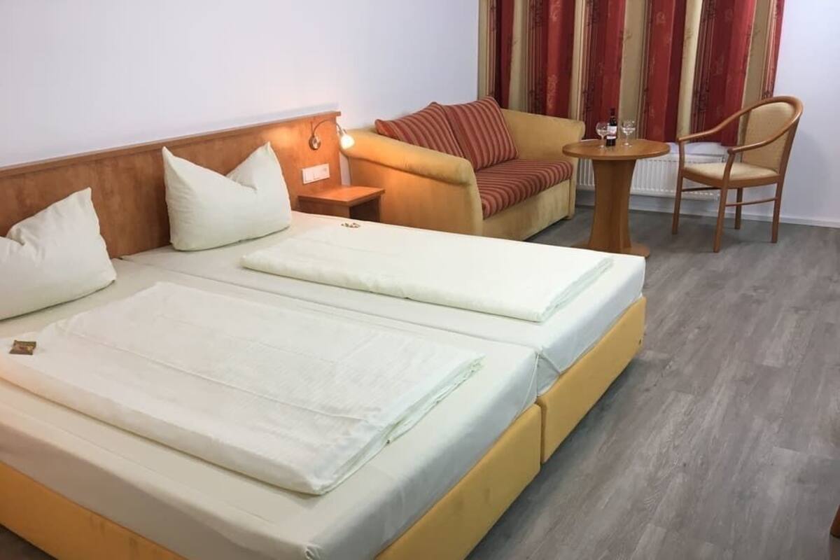 Ein Doppelbett mit Holz-Kopfteil, ein kleiner Nachttisch mit Lampe, ein Sofa mit gestreiften Kissen, ein runder Tisch mit Stuhl und ein Heizkörper.