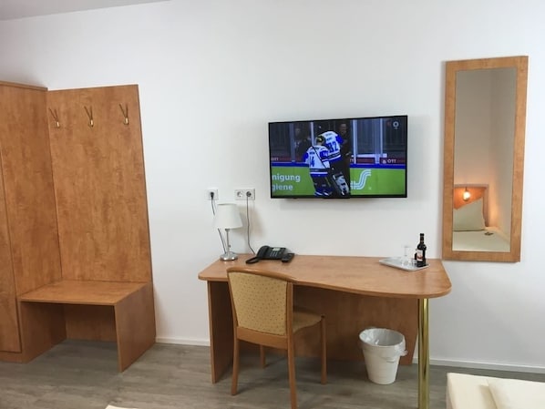 Una habitación con un escritorio de madera, una silla, un televisor montado en la pared, un espejo y un cubo de basura pequeño.