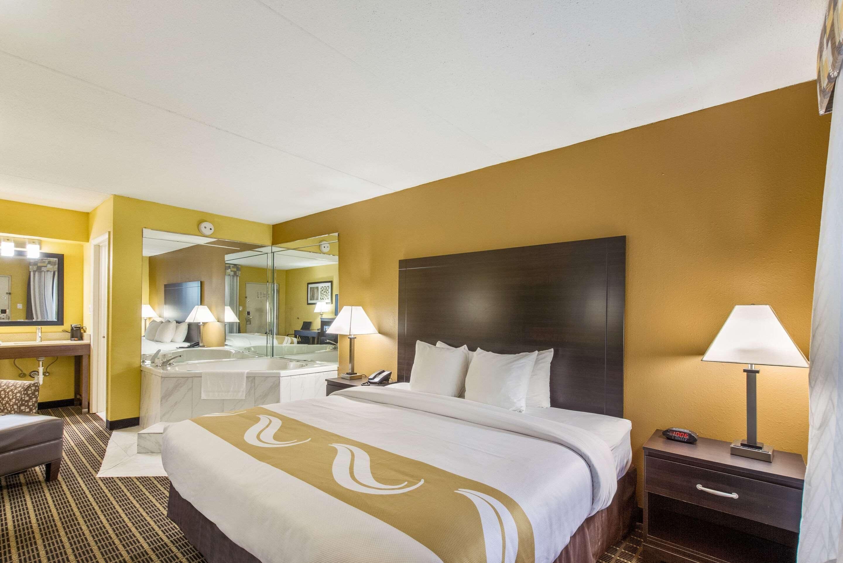 Hotelkamer met een groot bed, nachtkastje met een lamp, telefoon op een nachtkastje, spiegel en badkamer met badkuip en wastafel.