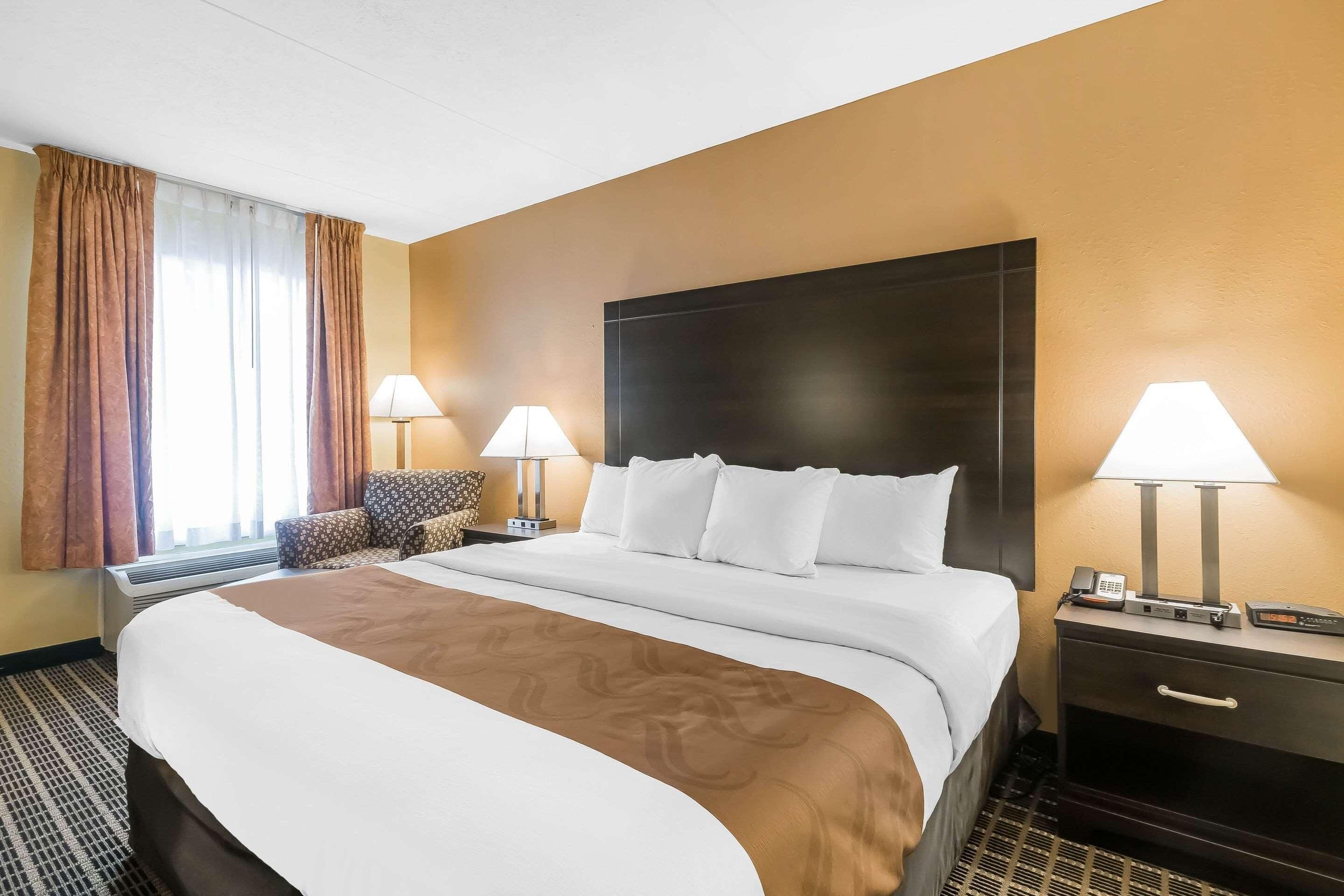 A hotel room with a large bed, two bedside lamps, a nightstand with a phone, and a chair.