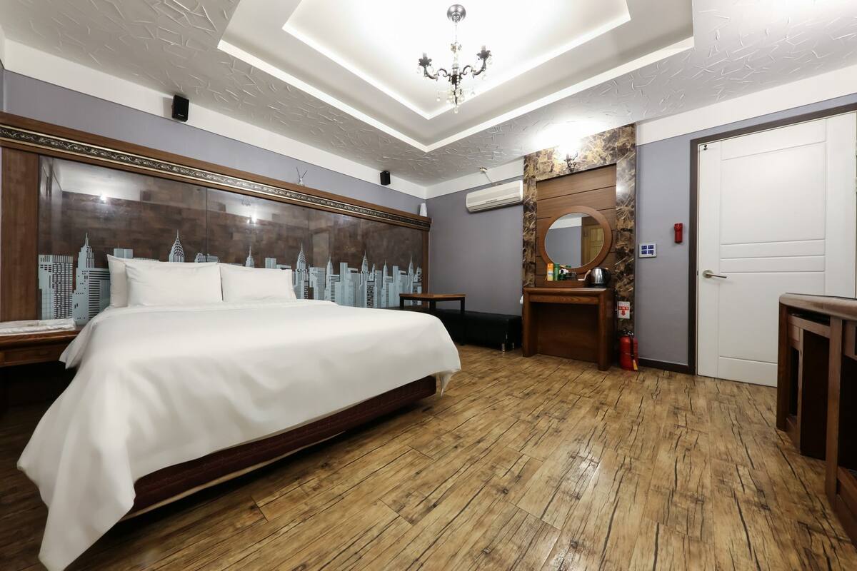 Una habitación de hotel con una cama grande, piso de madera, una obra de arte con una vista urbana en la pared, un espejo redondo y una puerta blanca.