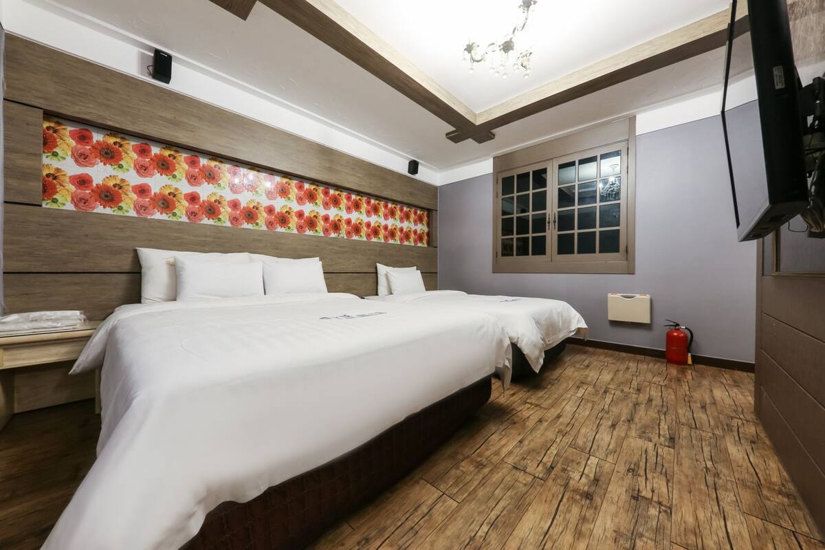 Un dormitorio con piso de madera, una cama grande con ropa de cama blanca, un cabecero estampado con motivos florales, una ventana con persianas, un televisor montado en la pared y un extintor.
