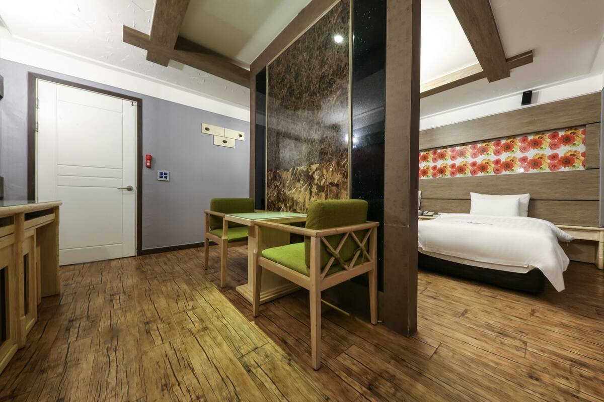 Habitación de hotel con piso de madera, cama con ropa blanca, una silla tapizada de verde, una mesita y un panel decorativo en la pared.