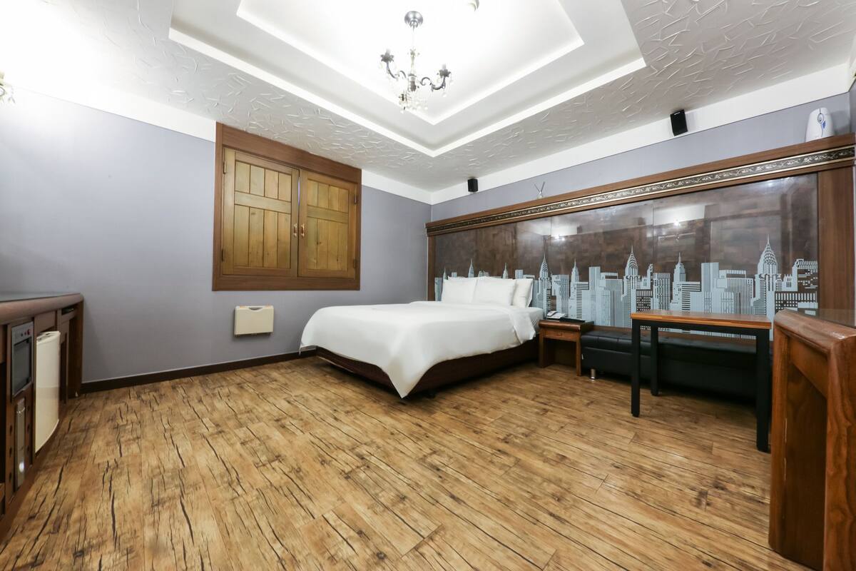Un dormitorio con piso de madera, una cama, una mesita de noche, un escritorio y un mural con vista a la ciudad en la pared.