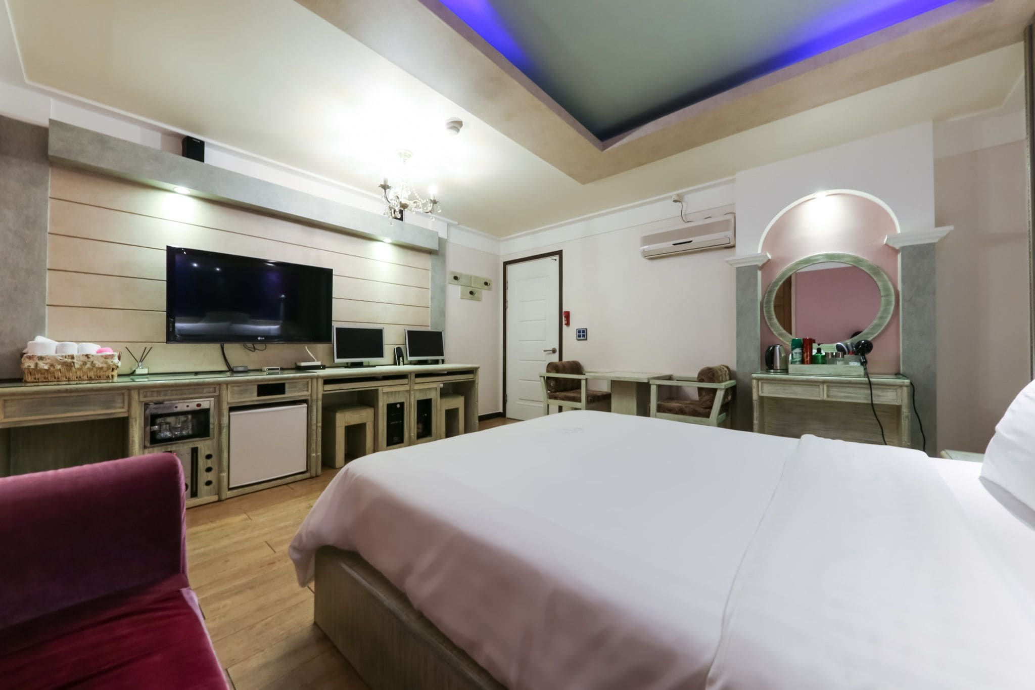 A hotel room with a large bed, a desk with a flat-screen TV, a kitchenette, a vanity with a mirror, and a ceiling with blue lighting.