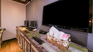 Une pièce dotée d’un grand téléviseur à écran plat fixé au mur, d’un bureau en bois avec deux écrans, un clavier et un panier rempli de mouchoirs.